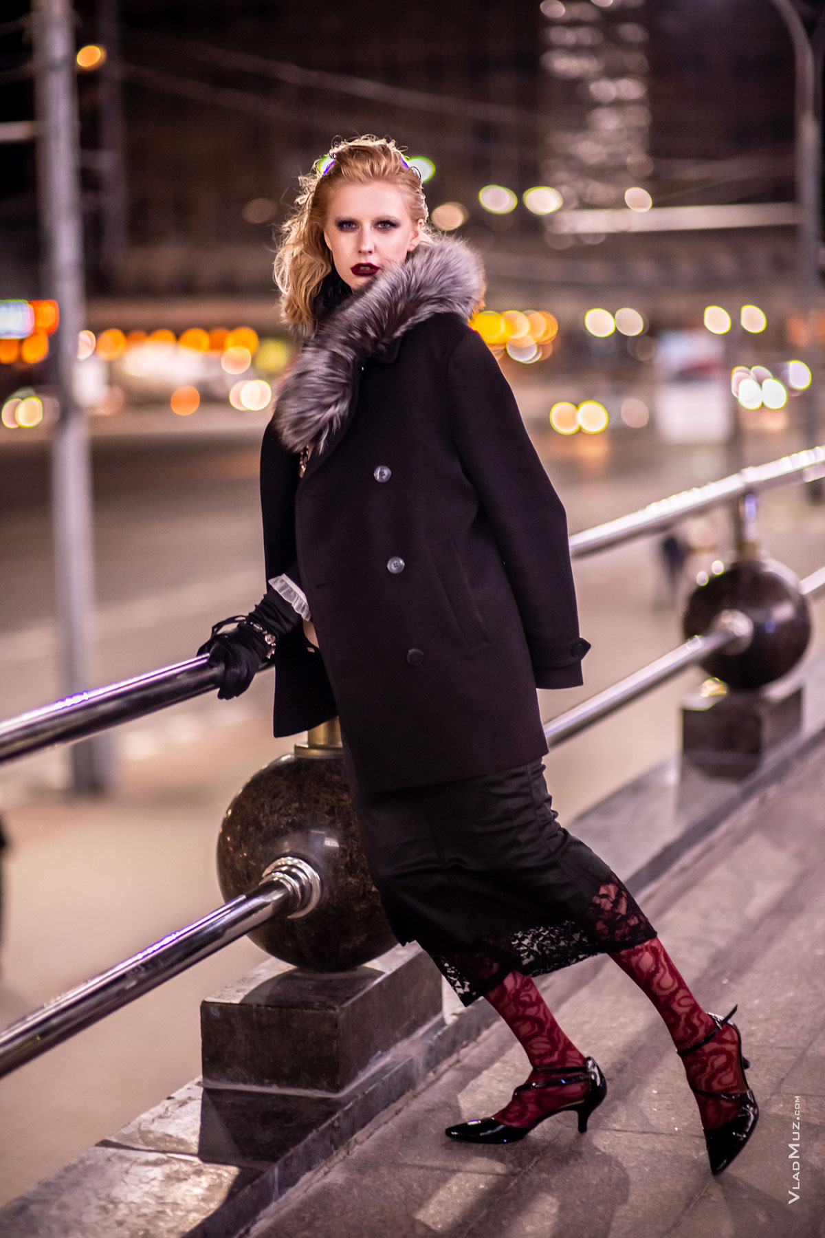 Фото девушки в полный рост в черном пальто с меховым воротником на фоне ночного города